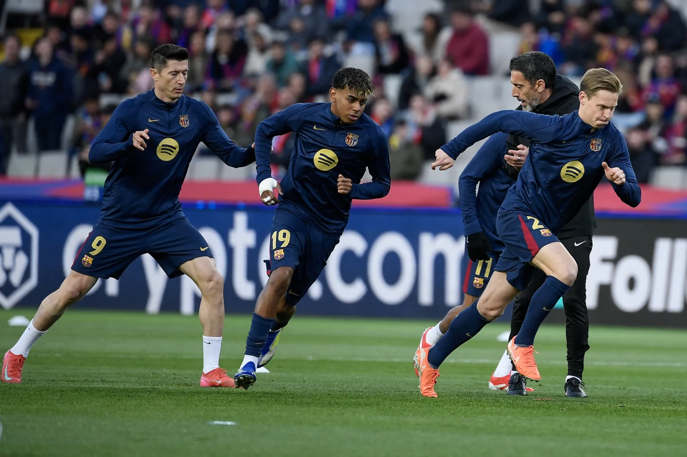 Robert Lewandowski, Lamine Yamal oraz Frenkie de Jong Czterech piłkarzy ubranych w niebieskie dresy wykonuje rozgrzewkę na stadionie przed rozpoczęciem meczu, skupiając się na ćwiczeniach biegowych na murawie.