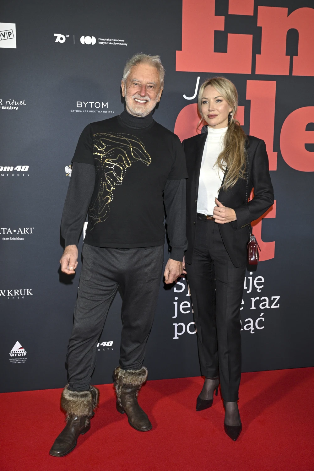 Rafał Olbiński z nową partnerką Dwie elegancko ubrane osoby stoją na czerwonym dywanie. Mężczyzna ma siwe włosy i brodę, ubrany jest w czarny sweter z motywem zwierzęcym oraz futrzane buty. Kobieta ma długie blond włosy, ubrana jest w czarny garnitur i białą koszulę. W tle widoczna j...