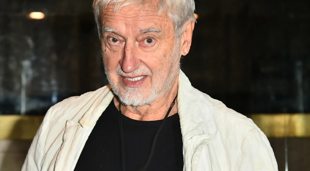 Rafał Olbiński Starszy mężczyzna z siwymi włosami i brodą, ubrany w czarną koszulkę oraz jasną kurtkę, stoi na tle ciemnego, nieostrego wnętrza.