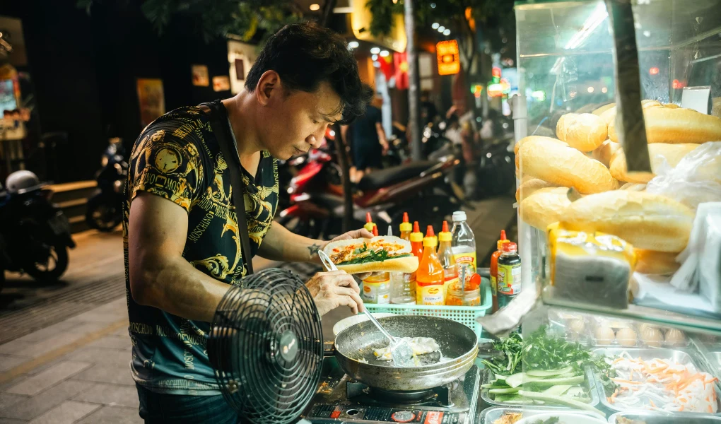 Mężczyzna przygotowuje jedzenie na ulicznym stoisku kuchni azjatyckiej, wokół widoczne składniki, sosy, warzywa i bagietki, w tle nocna sceneria miejskiej ulicy z motocyklami.