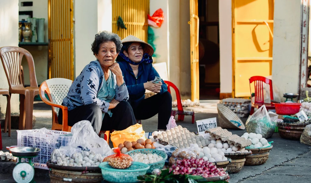 Dwie starsze kobiety siedzą przy stoiskach targowych z koszami wypełnionymi jajkami, warzywami i innymi produktami spożywczymi, otoczone jasnym światłem dnia oraz żółtymi i pomarańczowymi drzwiami w tle.