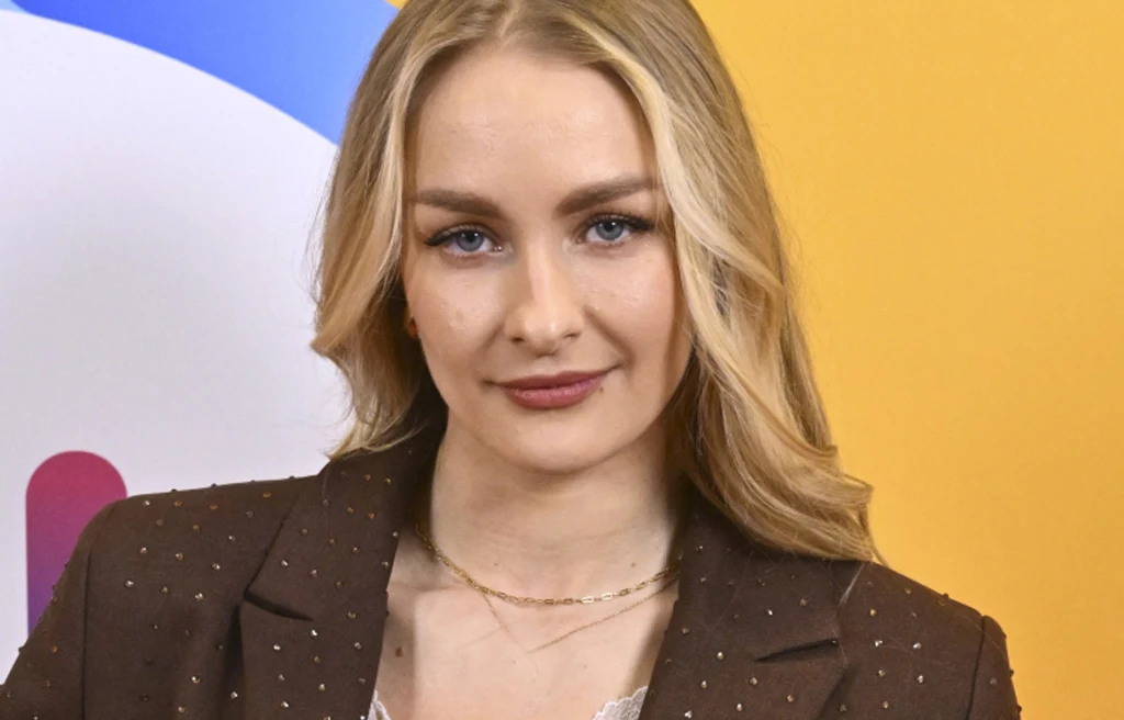 Sara Janicka Młoda kobieta o długich blond włosach w brązowej marynarce z delikatnym makijażem, stojąca na tle żółto-niebieskiej grafiki.