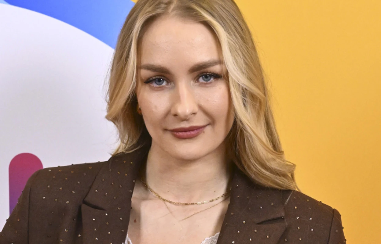 Sara Janicka Młoda kobieta o długich blond włosach w brązowej marynarce z delikatnym makijażem, stojąca na tle żółto-niebieskiej grafiki.