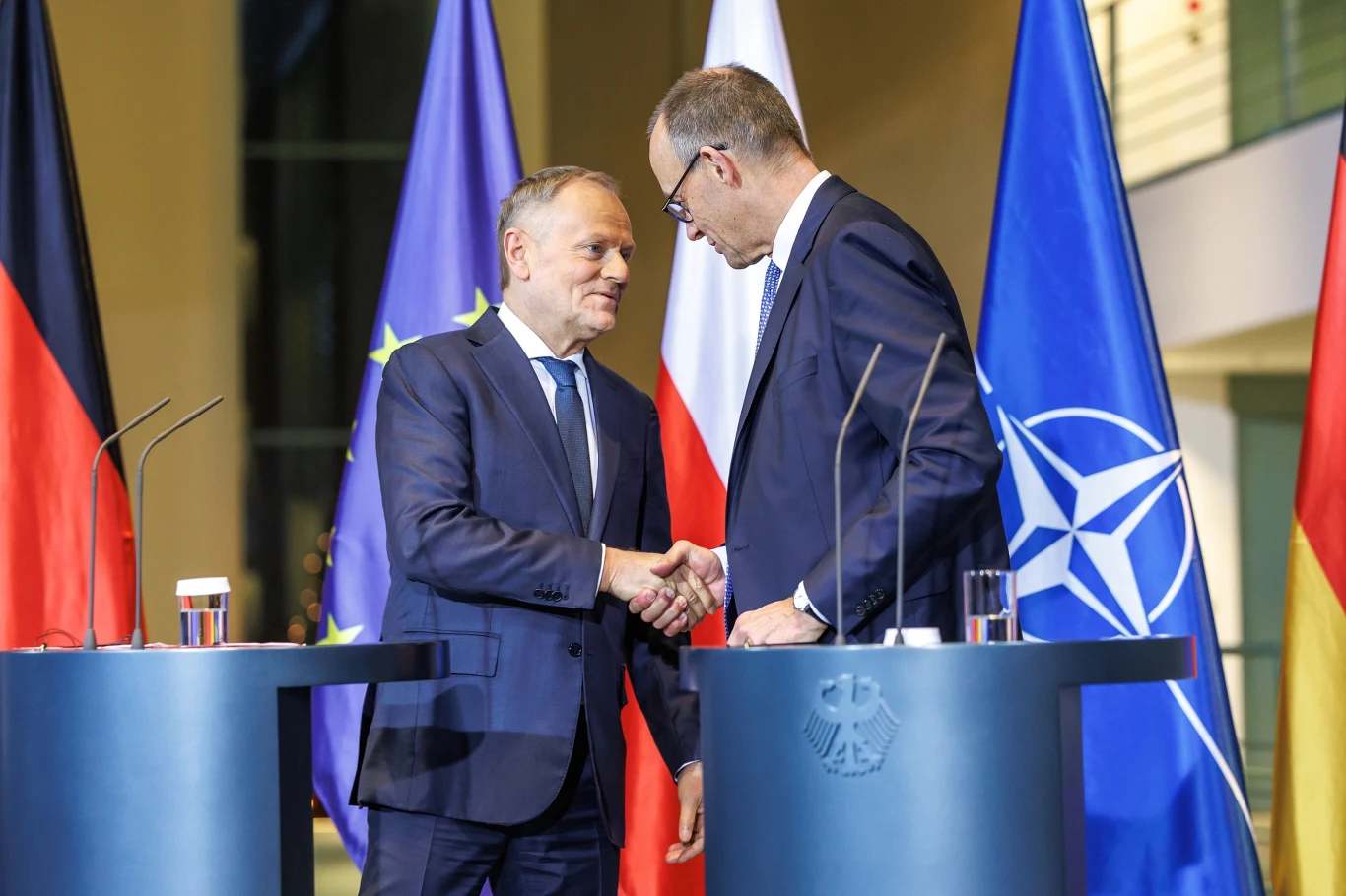 Donald Tusk na spotkaniu z Friedrichem Merzem Dwóch mężczyzn w garniturach podaje sobie ręce podczas oficjalnej konferencji prasowej, za nimi widoczne flagi Niemiec, Polski, Unii Europejskiej oraz NATO, a także mównice z mikrofonami.