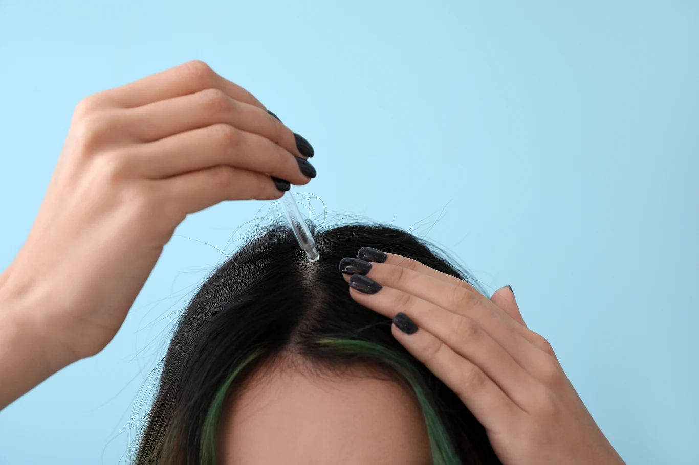 Nawilżające serum do skóry głowy to jeden ze sposobów radzenia sobie z przesuszonym skalpem Dłonie z ciemnym manicure nakładają płyn z pipety na przedziałek włosów ciemnowłosej osoby, tło jasnoniebieskie.