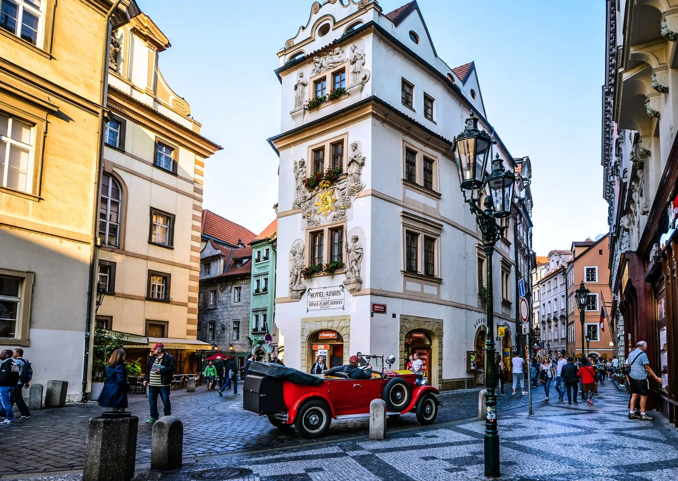 Zabytkowa kamienica z ozdobnymi rzeźbami na fasadzie, wokół niej brukowana uliczka i liczne stare budynki. Przed kamienicą stoi czerwony, zabytkowy samochód, a w tle spacerują ludzie podziwiający architekturę.