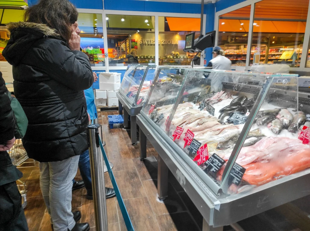 Stoisko rybne w supermarkecie z szerokim wyborem świeżych ryb i owoców morza, na pierwszym planie stojący przy ladzie klient wybierający produkt, wnętrze sklepu dobrze oświetlone.