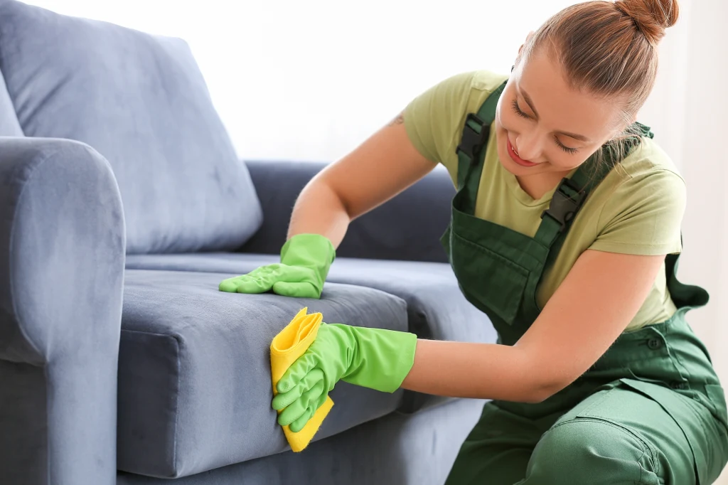 Zanim użyjesz środka czyszczącego, przetestuj go na małym fragmencie sofy Kobieta w zielonym stroju roboczym i rękawiczkach czyści niebieską sofę żółtą ściereczką, skupiając się na dokładnym czyszczeniu podłokietnika.
