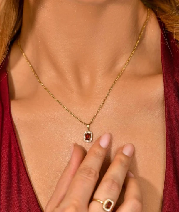 Luksusowe kosmetyki Złoty naszyjnik z prostokątnym wisiorkiem, ozdobionym czerwonym kamieniem i otoczonym drobnymi błyszczącymi kamieniami. Dłoń kobiety z pierścionkiem dopasowanym do naszyjnika delikatnie przytrzymuje wisior na tle bordowej sukienki.
