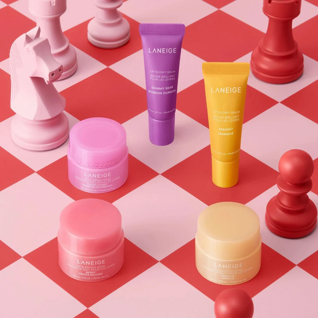Różnokolorowe kosmetyki Laneige w postaci kremów i balsamów ustawione na czerwono-białej szachownicy wśród różowych figur szachowych, dominują przyjemne pastelowe odcienie.