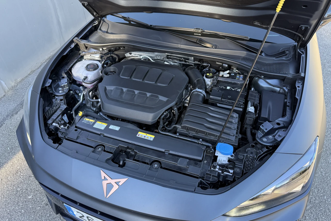 Silnik samochodowy umieszczony w komorze nowoczesnego pojazdu, widoczne elementy techniczne takie jak akumulator, przewody, zbiornik płynu chłodniczego oraz charakterystyczne logo producenta na pokrywie silnika.