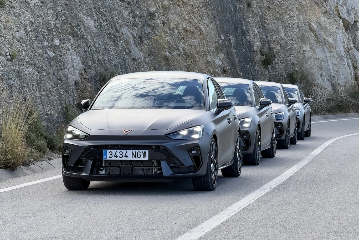 Cztery samochody sportowe marki Cupra ustawione jeden za drugim na krętej, asfaltowej drodze przy skalistej ścianie, widoczne z przodu, wszystkie w kolorze szarym.