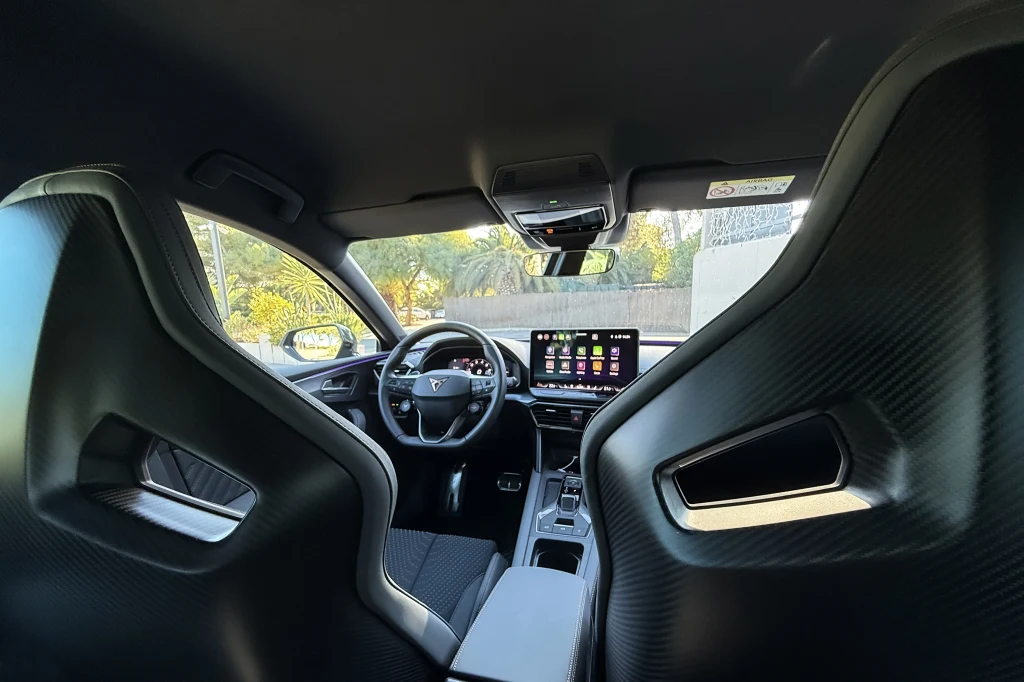 Wnętrze nowoczesnego samochodu sportowego, widok z tylnego siedzenia na deskę rozdzielczą, kierownicę, cyfrowy wyświetlacz oraz fotele kubełkowe wykończone włóknem węglowym.