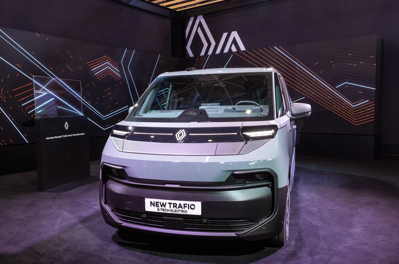 Czym wyróżnia się nowa klasa samochodów dostawczych? Nowoczesny, elektryczny samochód dostawczy Renault New Trafic prezentowany na eleganckim stoisku wystawienniczym, w otoczeniu futurystycznej scenografii z geometrycznymi motywami świetlnymi.