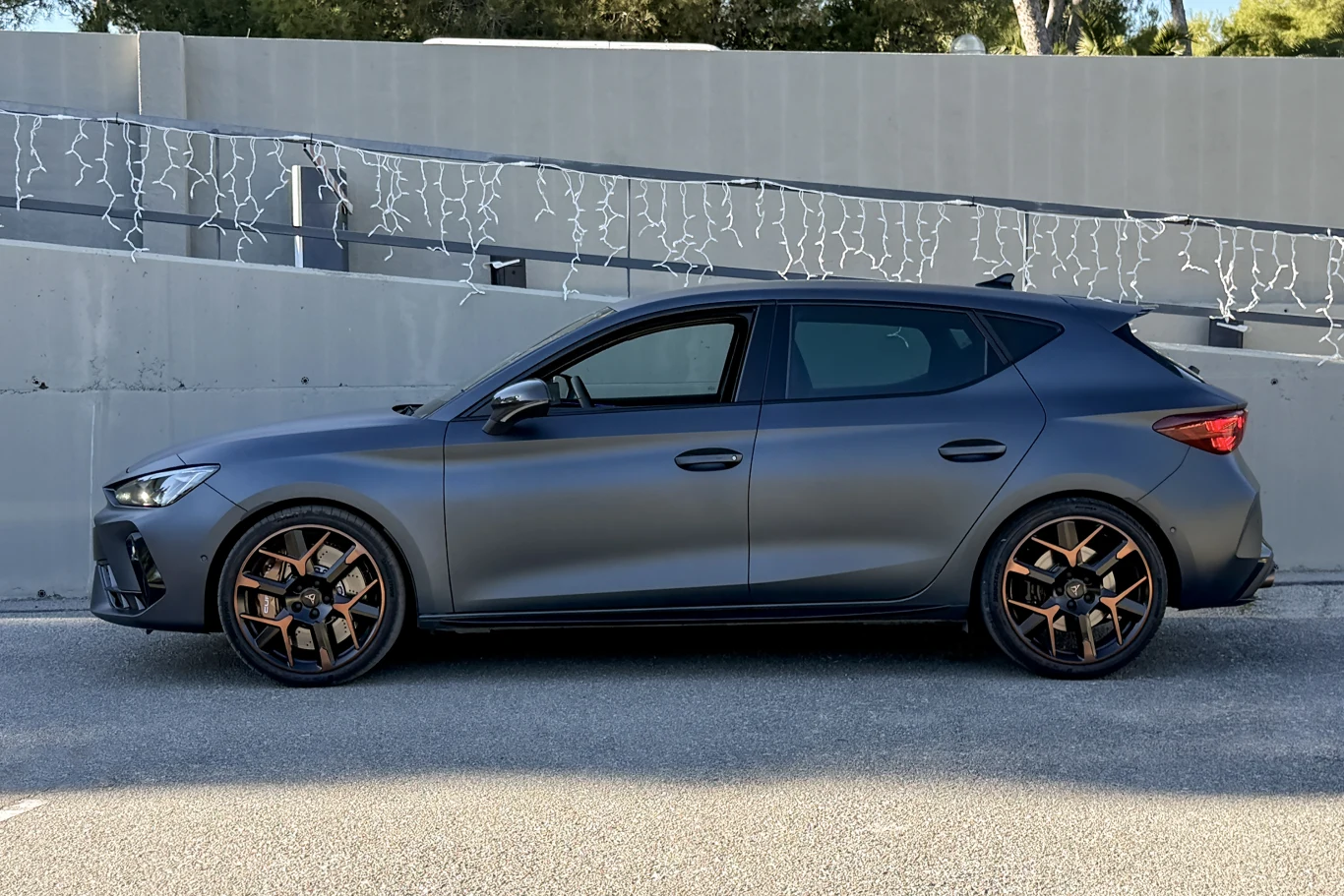 Ciemnoszary hatchback z przyciemnianymi szybami i sportowymi felgami stoi zaparkowany przy betonowej ścianie udekorowanej światełkami.