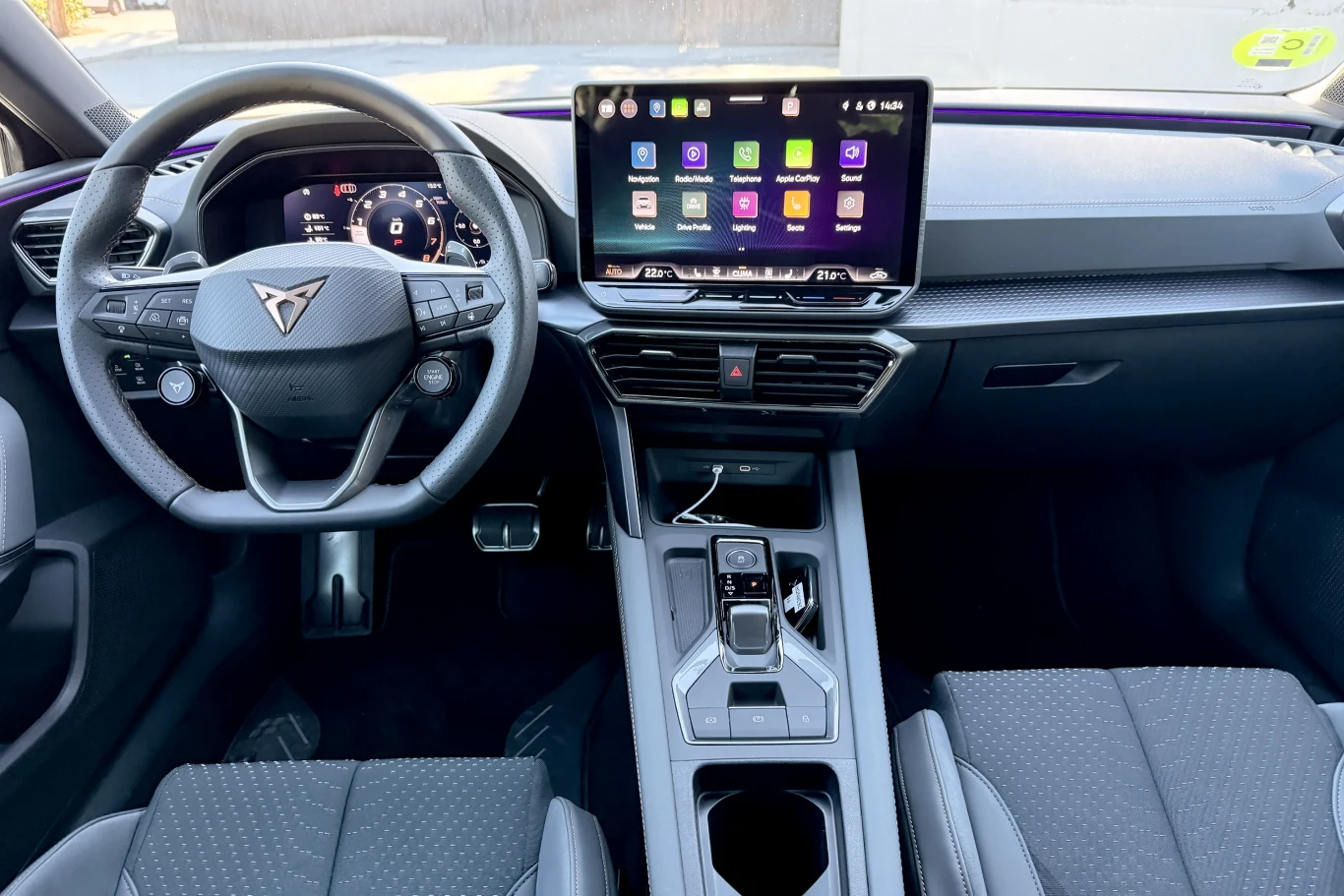 Nowoczesny kokpit samochodu z cyfrowym wyświetlaczem, dużym ekranem dotykowym z uruchomionymi aplikacjami oraz charakterystyczną kierownicą ozdobioną logo marki. Fotele przednie wykończone perforowaną tapicerką w ciemnym kolorze.