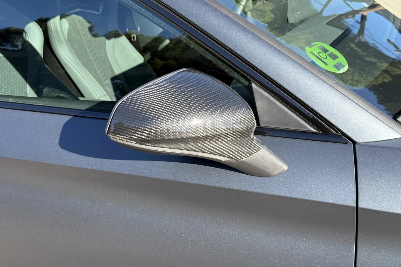 Detail lusterka bocznego z obudową z włókna węglowego w nowoczesnym samochodzie, widoczny fragment drzwi i wnętrza, na szybie zielona naklejka.