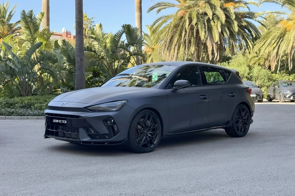 Czarny, sportowy hatchback o dynamicznej sylwetce stoi zaparkowany na tle tropikalnej roślinności i palm. Auto ma nowoczesne światła LED, duże felgi i agresywny wygląd.