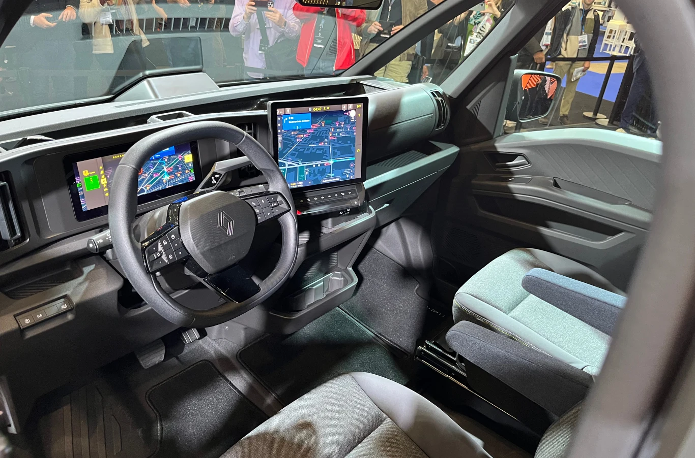 Na pokładzie dostępny jest Asystent Google, dzięki któremu kierowca może głosowo zmieniać ustawienia, robić notatki czy wywoływać informacje z sieci. Wnętrze nowoczesnego samochodu ciężarowego z cyfrowymi wyświetlaczami na desce rozdzielczej, dużym ekranem centralnym pokazującym mapę, ergonomicznie ukształtowanymi fotelami i kierownicą, wokół widoczni są ludzie przyglądający się pojazdowi.