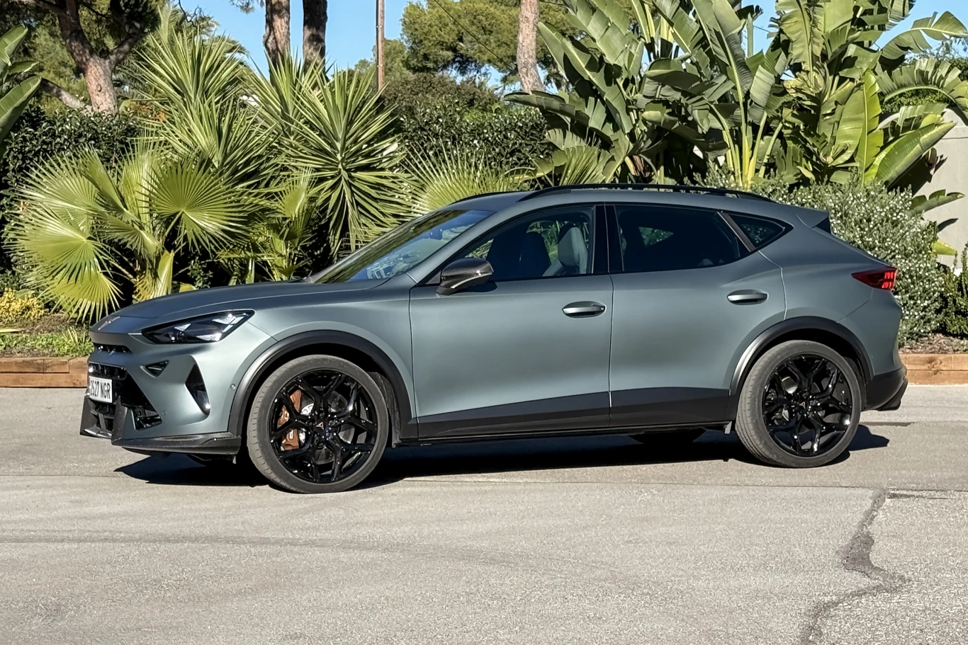 Nowoczesny, matowoszary samochód typu SUV o sportowej sylwetce stoi zaparkowany na tle bujnych, egzotycznych roślin i palm w słoneczny dzień.