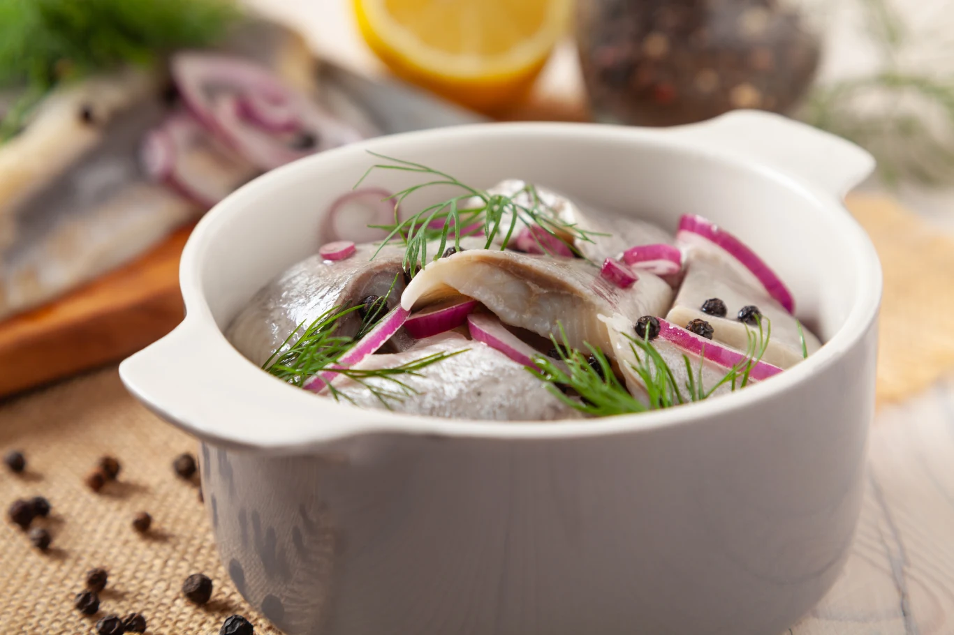 Śledzie to obowiązkowy element świątecznego menu Porcja śledzi w zalewie z cebulą czerwoną i gałązkami koperku ułożona w białym naczyniu, w tle plasterek cytryny i ziarnka czarnego pieprzu.