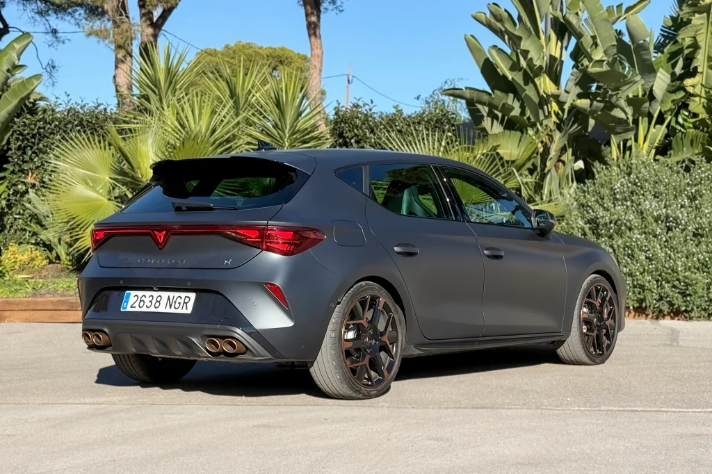 Szary samochód typu hatchback z ciemnymi felgami stoi na parkingu otoczonym egzotyczną roślinnością. W tle widać drzewa oraz błękitne niebo. Tablica rejestracyjna z numerem 2638 NGR umieszczona jest z tyłu auta.