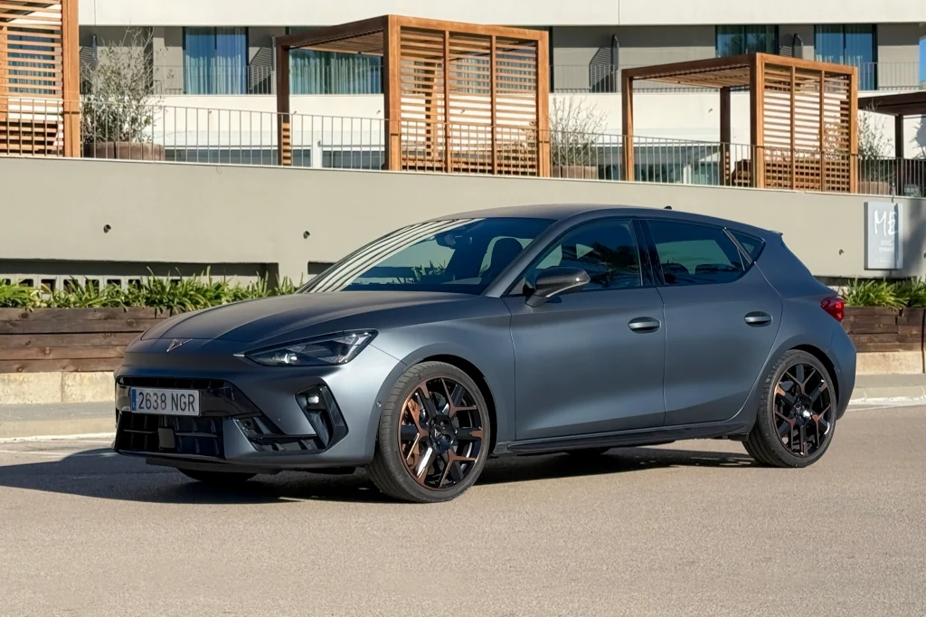 Nowoczesny, sportowy samochód klasy hatchback w matowym, grafitowym kolorze stoi zaparkowany na tle budynku z drewnianymi elementami architektonicznymi. Auto wyróżnia się czarnymi felgami i dynamicznymi liniami nadwozia.