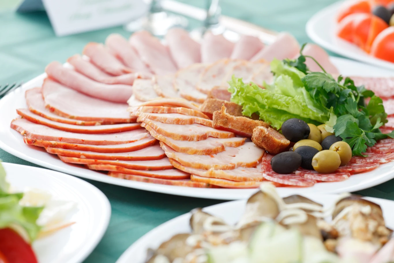 Talerz różnorodnych wędlin w plastrach, ułożonych promieniście, z dodatkiem sałaty, zielonych i czarnych oliwek oraz kawałkach pieczywa, na stole wśród innych potraw.