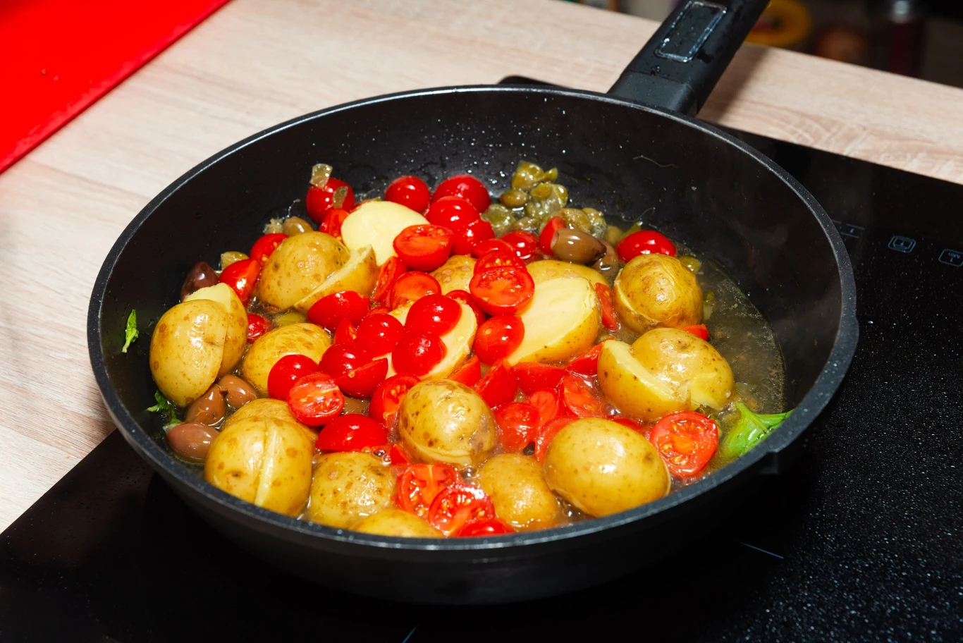 Kolorowa mieszanka młodych ziemniaków, pomidorków koktajlowych i czerwonych porzeczek smażona na patelni, widoczna na kuchennej płycie grzewczej, otoczona kawałkami oliwek i papryki.