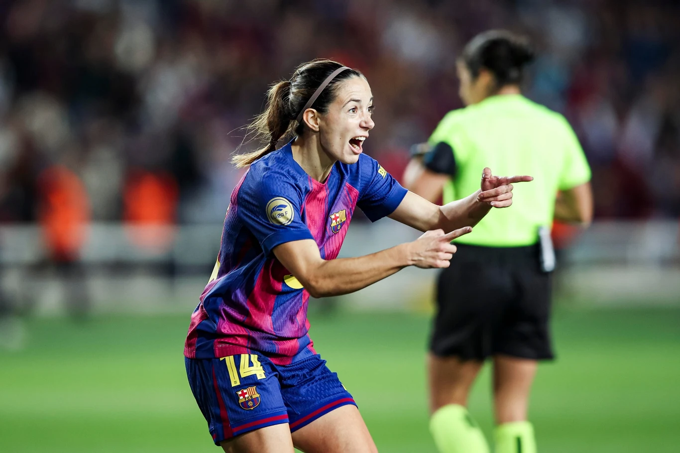 Piłkarka w stroju FC Barcelony z numerem 14 na plecach świętuje z uśmiechem na twarzy, gestykulując obiema rękami podczas meczu. W tle widoczna sędzia oraz rozmyte sylwetki innych uczestników spotkania.
