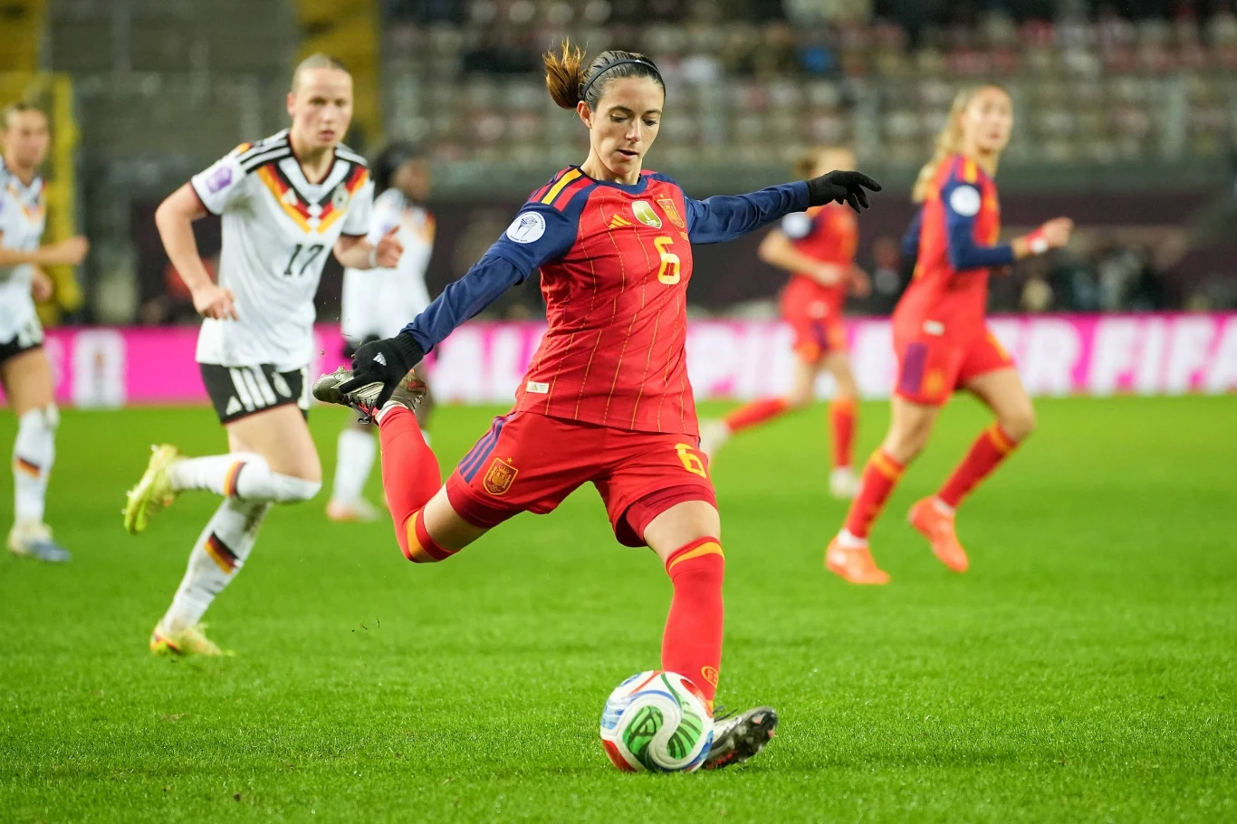 Piłkarka w czerwonym stroju podczas dynamicznej akcji kopnięcia piłki na boisku, wokół niej zawodniczki drużyny przeciwnej ubrane w białe stroje, w tle widać trybuny z widownią.