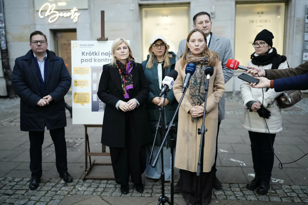 Grupa sześciu osób stojąca przed tablicą z informacjami podczas konferencji prasowej, przy kilku mikrofonach, na tle budynku miejskiego. Jedna z kobiet przemawia, reszta słucha lub czeka na swoją kolej.