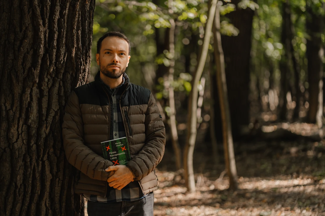 Marek Józefiak Mężczyzna w jesiennej kurtce oparty o pień drzewa w lesie, trzymający książkę z zieloną okładką, w tle gęste drzewa i rozproszone światło słoneczne.
