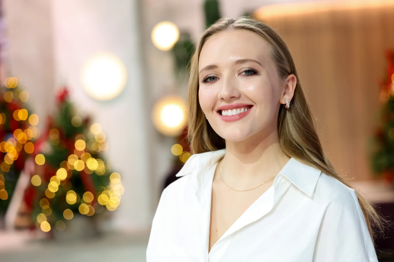 Karolina Matej jest wnuczką Grażyny Szapołowskiej Młoda kobieta z długimi blond włosami uśmiecha się, ubrana w białą koszulę, w tle rozmyte światełka świąteczne oraz ozdobione choinki.