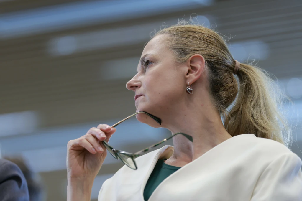 Kobieta z blond włosami spiętymi w kucyk, ubrana w jasny żakiet, trzyma okulary w dłoni i patrzy w bok w zamyślonym wyrazie twarzy. Tło jest rozmyte, skupiając uwagę na postaci.