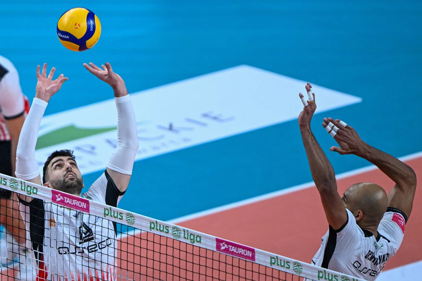 Dwóch siatkarzy w trakcie meczu, jeden z nich wyskakuje do wystawienia piłki, drugi przygotowuje się do ataku. Kolorowa piłka unosi się tuż nad siatką, a na parkiecie widoczne są logotypy sponsorów.