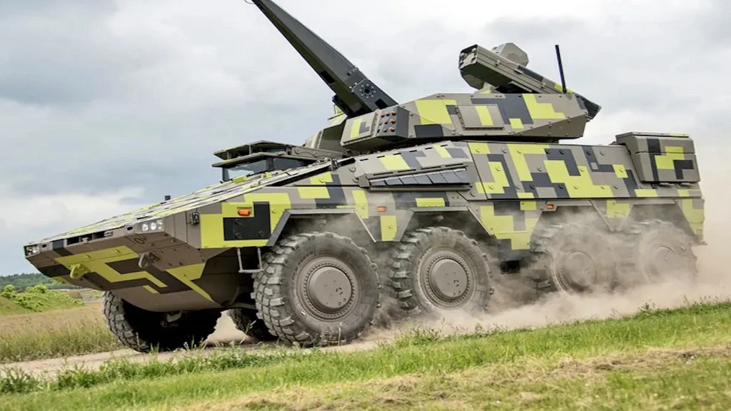 Niemcy zmieniają fabryki samochodów w zakłady produkcji broni Nowoczesny, opancerzony pojazd wojskowy na sześciu dużych kołach w ruchu po trawiastej drodze. Pojazd pokryty jest wzorem maskującym w odcieniach zieleni, szarości i czerni oraz wyposażony w stanowisko ogniowe z zamontowaną bronią.