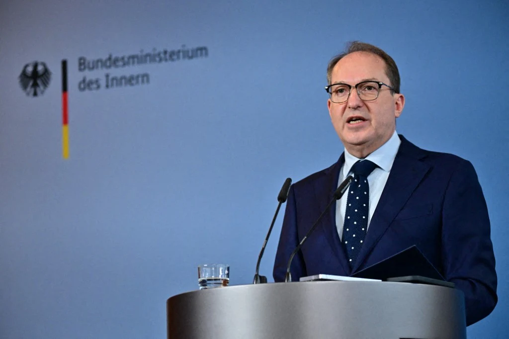 Niemiecki minister spraw wewnętrznych Alexander Dobrindt Mężczyzna w garniturze i okularach przemawia przy mównicy, z tyłu widoczne logo niemieckiego Ministerstwa Spraw Wewnętrznych na niebieskiej ścianie. Przed nim mikrofony, szklanka wody oraz teczka z dokumentami.