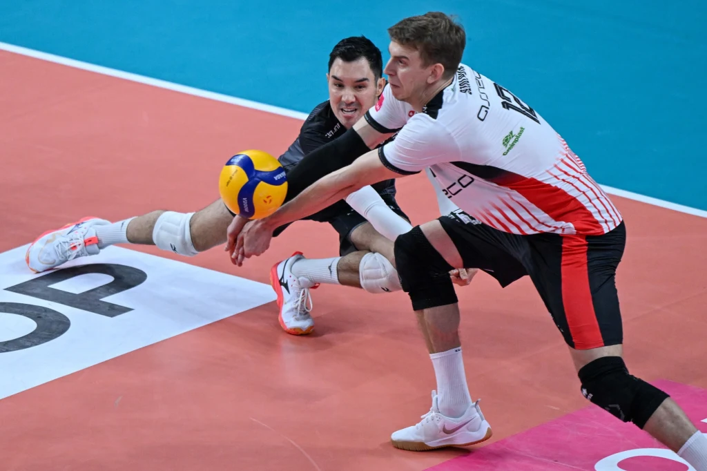 Erik Shoji (z lewej) i Artur Szalpuk, siatkarze Asseco Resovii Rzeszów, która zagra w Lidze Mistrzów dzięki dzikiej karcie Dwóch siatkarzy w trakcie dynamicznej akcji odbioru piłki, jeden w białym stroju z czerwonymi elementami, drugi w czarnym, obaj skoncentrowani na grze, na tle kolorowego boiska.