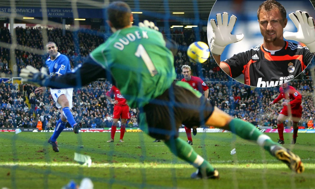 Piłkarz ubrany w niebieski strój wykonuje rzut karny, naprzeciwko niego bramkarz w zielonym stroju z nazwiskiem 'Dudek' na plecach rzuca się w lewą stronę bramki podczas obrony. W tle trybuny wypełnione kibicami, na boisku widać też innych zawodników. ...