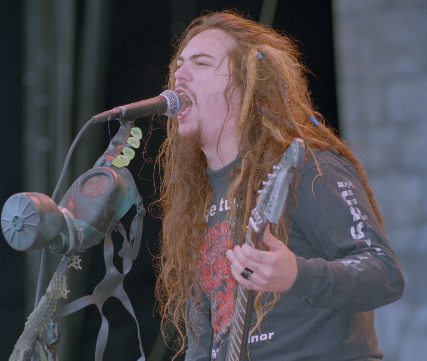 Max Cavalera w składzie Sepultury z okresu promocji płyty "Chaos A.D." Młody mężczyzna z długimi, splątanymi włosami wykonuje utwór na żywo, śpiewając do mikrofonu i grając na gitarze elektrycznej. W tle widoczna maska przeciwgazowa zawieszona na statywie mikrofonu, artysta ma intensywny wyraz twarzy i ubranie z charakter...
