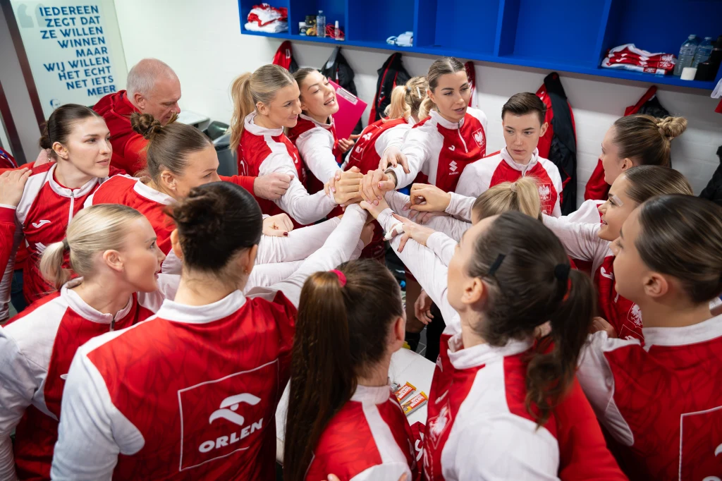 Drużyna kobiet ubrana w czerwono-białe stroje sportowe zbiera się w szatni, tworząc okrąg i łącząc ręce na znak jedności oraz wsparcia przed wyjściem na boisko. W tle widoczne niebieskie szafki oraz napisy motywacyjne na ścianie.