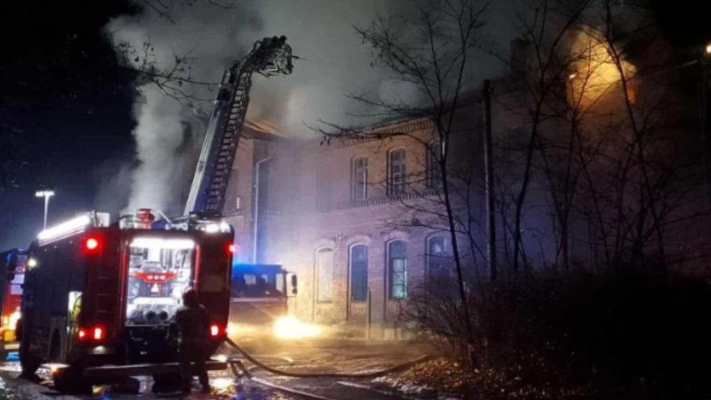 Płonący budynek, z którego wydobywa się gęsty dym, oświetlony jest światłami pojazdów strażackich oraz reflektorami. Na pierwszym planie znajduje się wóz strażacki z rozłożoną drabiną oraz pracujący strażacy.