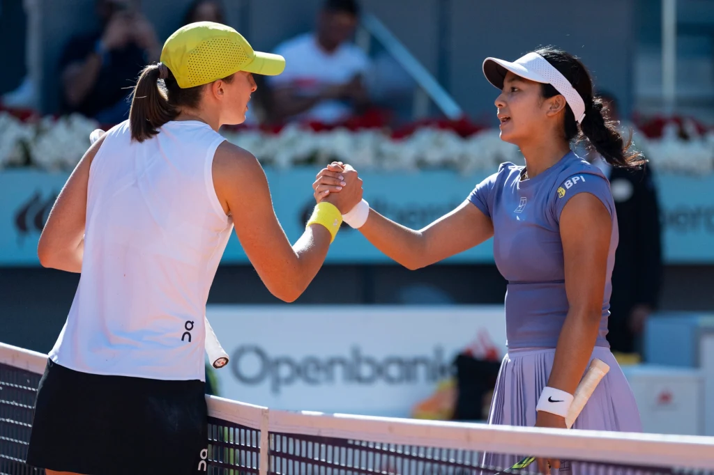 Dwóch tenisistki podają sobie ręce nad siatką na korcie tenisowym po zakończonym meczu, obie ubrane w stroje sportowe, w tle widoczni kibice oraz ogrodzenie kortu.