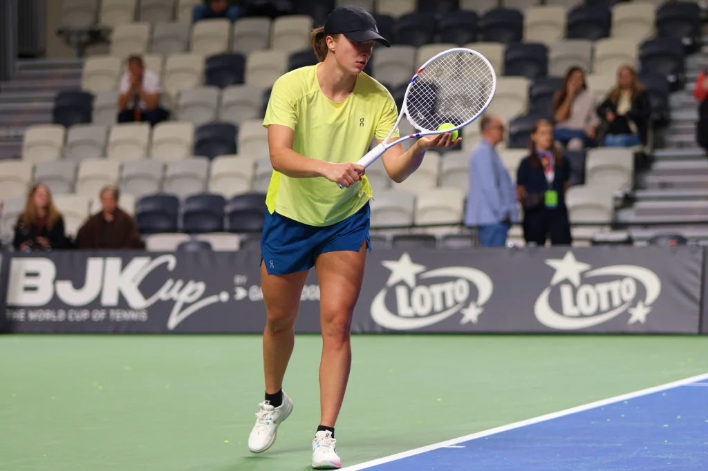 Tenisistka podczas meczu przygotowuje się do odbioru piłki na korcie w hali. W tle widoczne są puste krzesła oraz pojedynczy widzowie na trybunach, a także banery sponsorskie i baner turnieju BJK Cup.