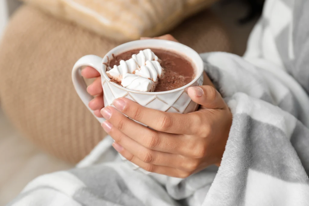 Dwie dłonie trzymają kubek gorącej czekolady z bitą śmietaną, osoba owinięta jest w szary koc na tle przytulnego wnętrza.