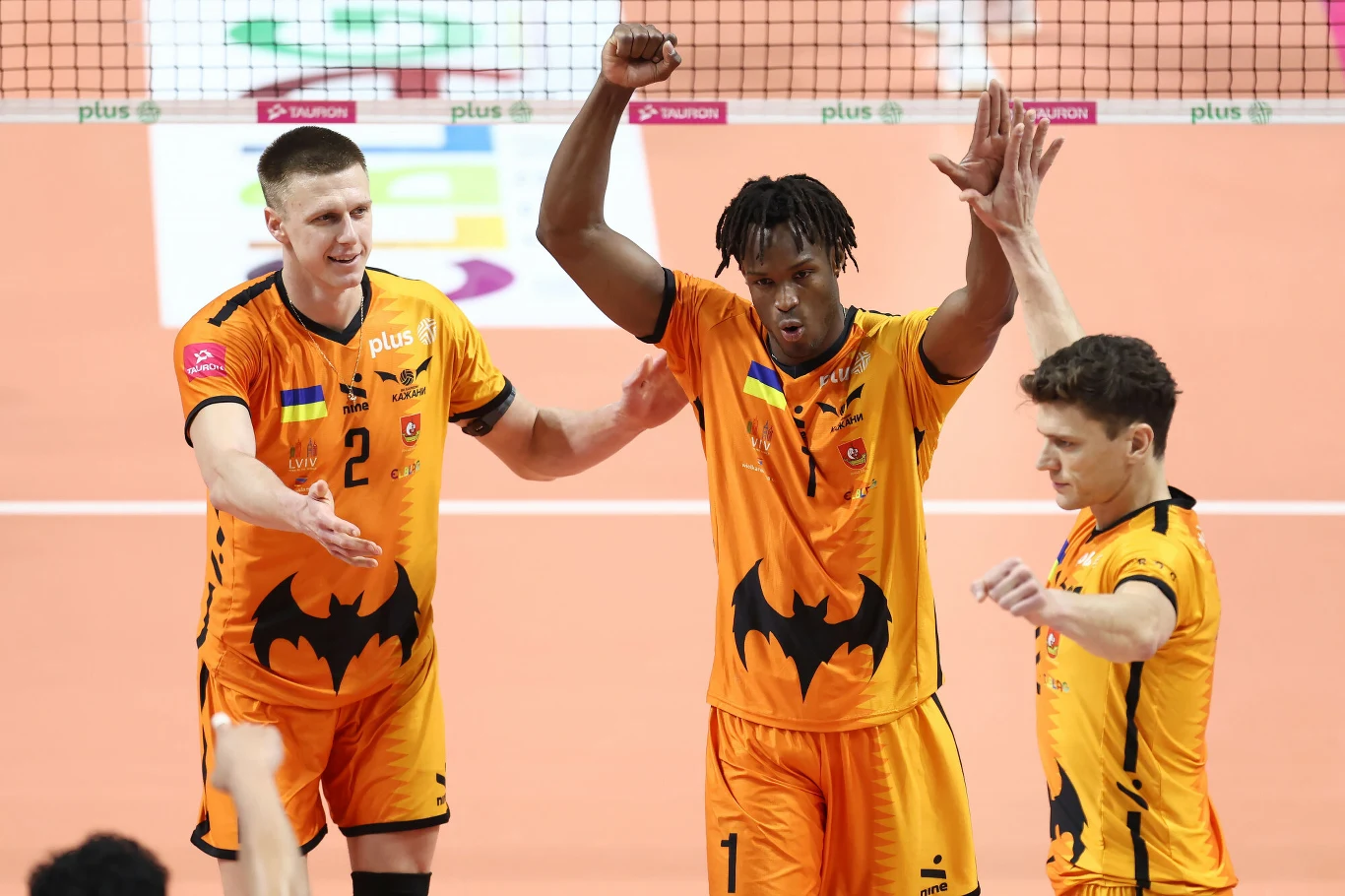 Siatkarze Barkomu Każany Lwów znów mogli świętować Trzech siatkarzy ubranych w pomarańczowe stroje z czarnym logo nietoperza stoi na boisku, jeden z nich unosi ręce w geście triumfu, pozostali patrzą w jego stronę, w tle widoczna siatka i fragmenty hali sportowej.