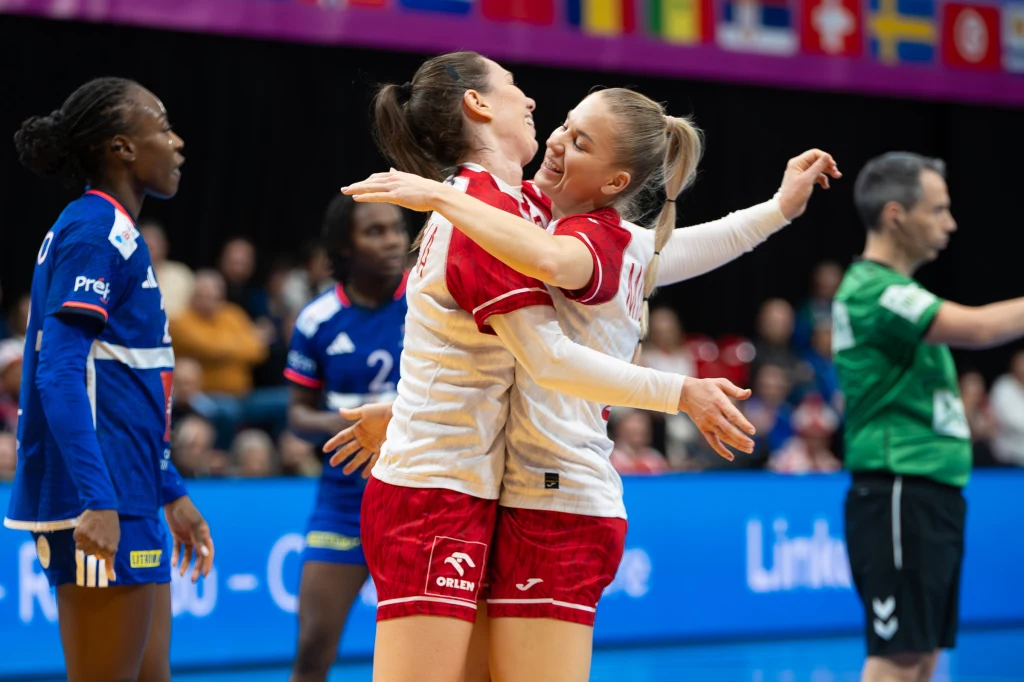 Monika Kobylińska i Daria Michalak miały się z czego cieszyć w pierwszej połowie meczu z Francją Dwie zawodniczki drużyny w białych koszulkach i czerwonych spodenkach cieszą się po udanej akcji, obejmując się na boisku do piłki ręcznej. W tle zawodniczki drużyny przeciwnej w niebieskich strojach oraz sędzia. Za nimi widoczne są flagi różnych państw.