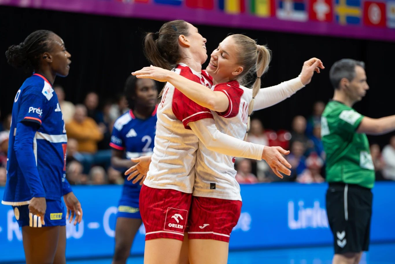 Monika Kobylińska i Daria Michalak miały się z czego cieszyć w pierwszej połowie meczu z Francją Dwie zawodniczki drużyny w białych koszulkach i czerwonych spodenkach cieszą się po udanej akcji, obejmując się na boisku do piłki ręcznej. W tle zawodniczki drużyny przeciwnej w niebieskich strojach oraz sędzia. Za nimi widoczne są flagi różnych państw.