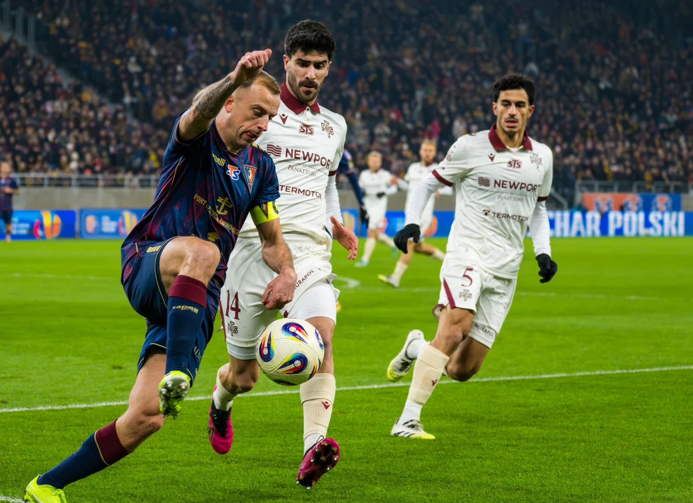 Intensywna rywalizacja trzech piłkarzy podczas meczu piłkarskiego na stadionie, zawodnik w ciemnym stroju próbuje przejąć piłkę, podczas gdy dwóch piłkarzy rywalizującej drużyny w białych strojach broni dostępu do bramki, w tle widać zapełnione trybuny.